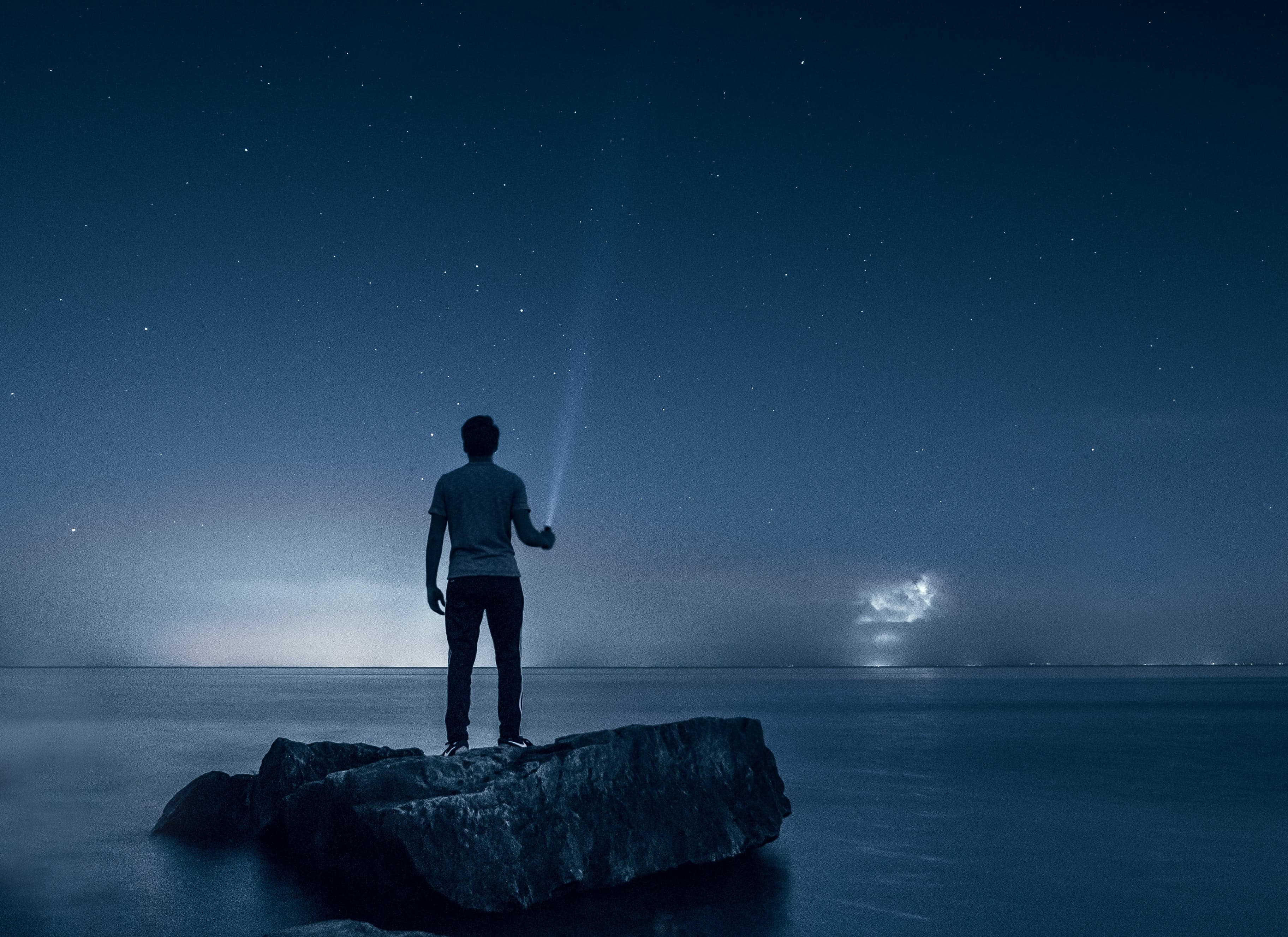 A person standing on a rock in the night, which is sorounded by water. The person is holding a pocket lamp to search the night sky.